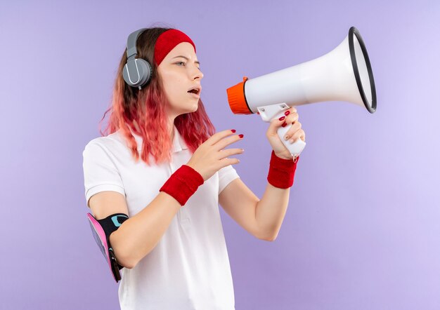 Young sporty woman shouting to megaphone loking aside standing over purple wall