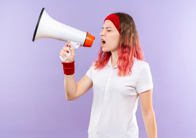 Молодая спортивная женщина кричит в мегафон с агрессивным выражением лица, стоя над фиолетовой стеной