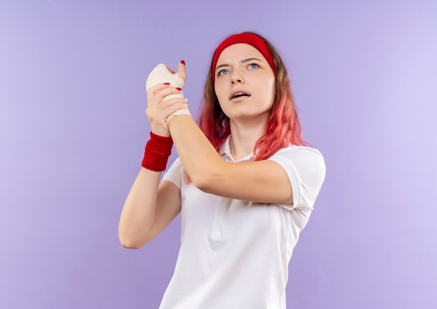 Giovane donna sportiva che tocca il suo polso bendato sensazione di dolore alzando lo sguardo in piedi sopra la parete viola