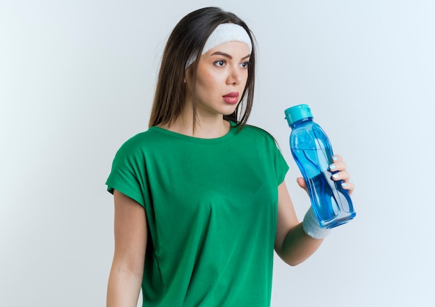 Бесплатное фото Молодая спортивная женщина с повязкой на голову и браслетами, держащая бутылку с водой, смотрит прямо