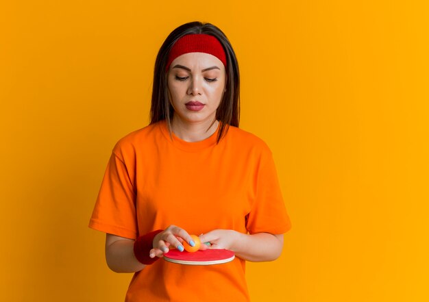 Young sporty woman wearing headband and wristbands holding and looking at ping pong racket with ball on it touching ball 