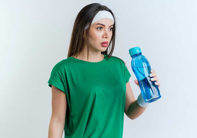 Молодая спортивная женщина с повязкой на голову и браслетами, держащая бутылку с водой, смотрит прямо