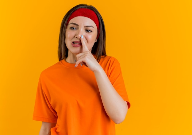 Young sporty woman wearing headband and wristbands looking at side putting hand on nose 