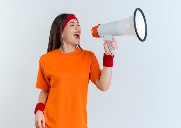 Young sporty woman wearing headband and wristbands turning head to side shouting in loud speaker with closed eyes isolated