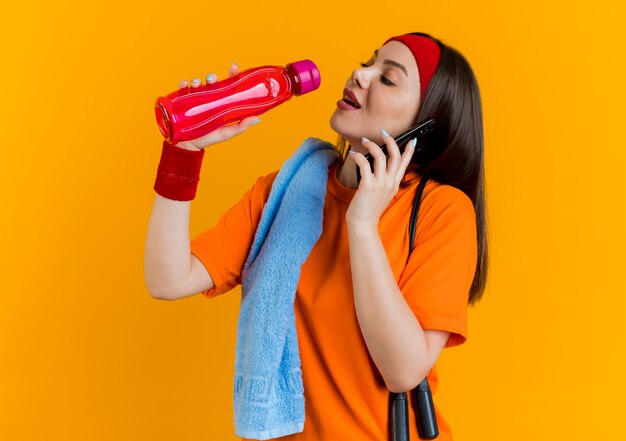 병을 들고 물을 마시려고 전화로 이야기하는 어깨에 점프 로프와 수건으로 머리띠와 팔찌를 착용하는 젊은 스포티 한 여자