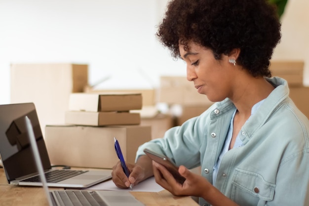 Young startup owner holding smartphone and checking order details. Attractive Black woman working on computer surrounded by cardboard parcels and making notes. Business, e-commerce concept