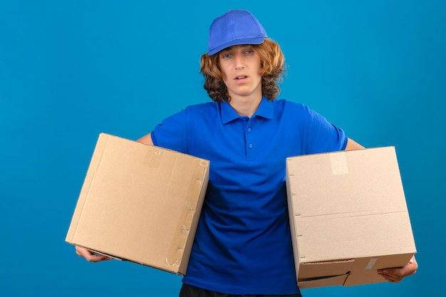 Молодой переутомленный доставщик в синей рубашке поло и кепке с картонными коробками выглядит усталым, стоя на изолированном синем фоне