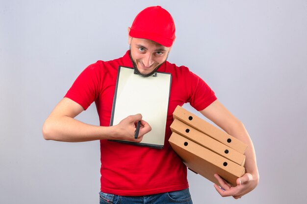 격리 된 흰색 배경 위에 서명을 요구하는 클립 보드를 들고 피자 상자의 스택과 함께 빨간색 폴로 셔츠와 모자를 입고 긴장 배달 남자 이상 영
