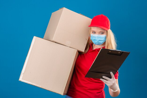 Young over strained delivery woman wearing red polo shirt and cap in medical protective mask standing with cardboard boxes while holding clipboard in other hand looking at it over isolated blue