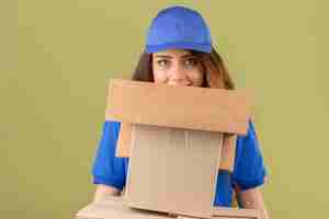 Foto gratuita giovane donna di consegna tesa con sguardo sornione che indossa polo blu e cappello in piedi con una pila di pacchi sorridente su sfondo verde isolato