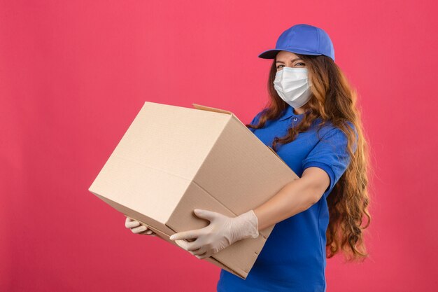 Young over strained displeased delivery woman with curly hair wearing blue polo shirt and cap in medical protective mask standing with large box over isolated pink background