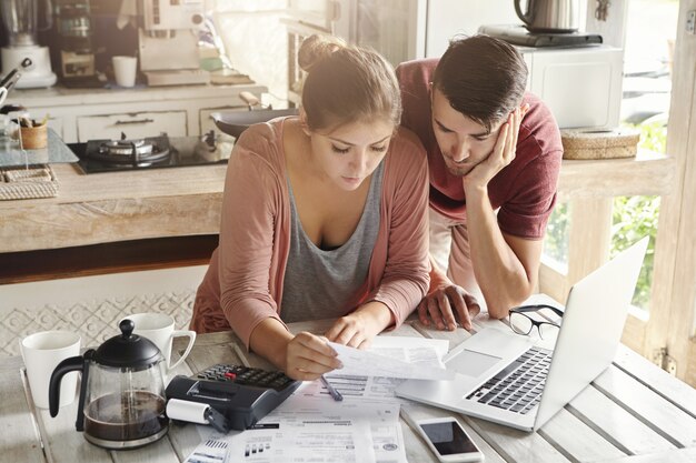 Young stressed family paying utility bills online using laptop. Worried woman holding document, calculating domestic expenses together with her husband