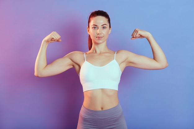 Young strong woman looking directly at camera, raising both hands and showing her biceps, wearing top and leggins, isolated over color background, lady with perfect body and ponytail.