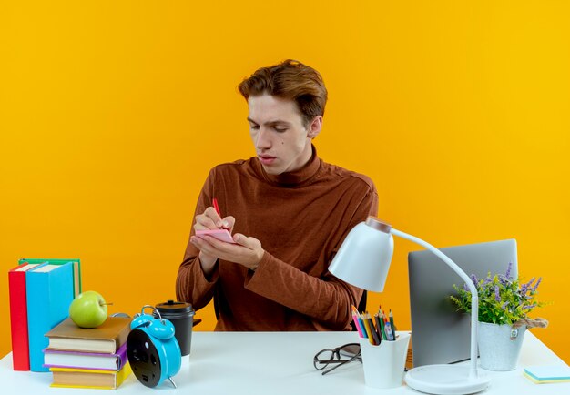 Young student boy sitting at desk with school tools writing something isolated on yellow wall