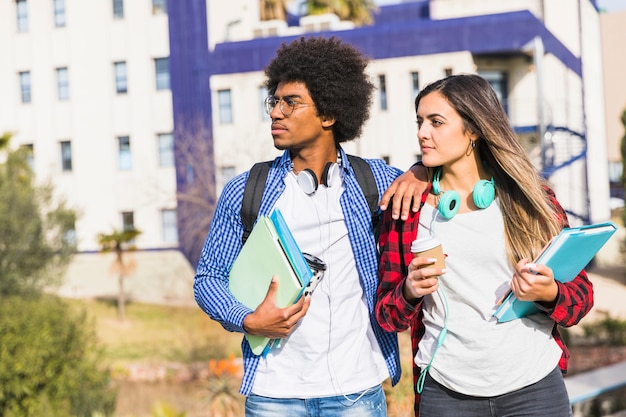 Young student couple holding books and disposable coffee cup standing against campus looking away