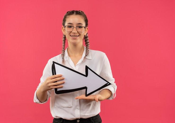 Free Photo young student girl in glasses with pigtails in white shirt holding white arrow looking to the front smiling cheerfully standing over pink wall