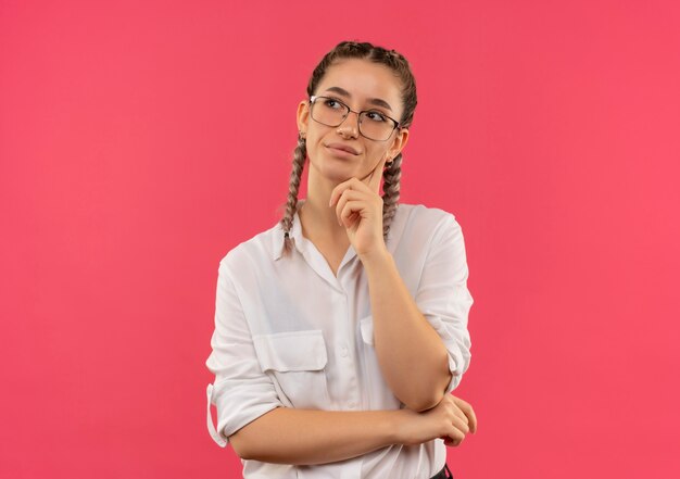 Young student girl in glasses with pigtails in white shirt looking aside with hand on chin puzzled standing over pink wall