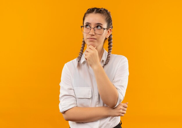 Young student girl in glasses with pigtails in white shirt looking aside with hand on chin with pensive expression on face thinking standing over orange wall