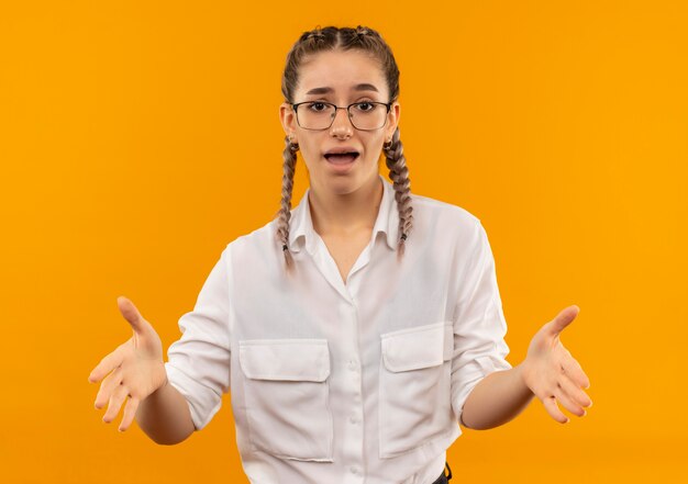 Young student girl in glasses with pigtails in white shirt looking confused and very anxious standing over orange wall