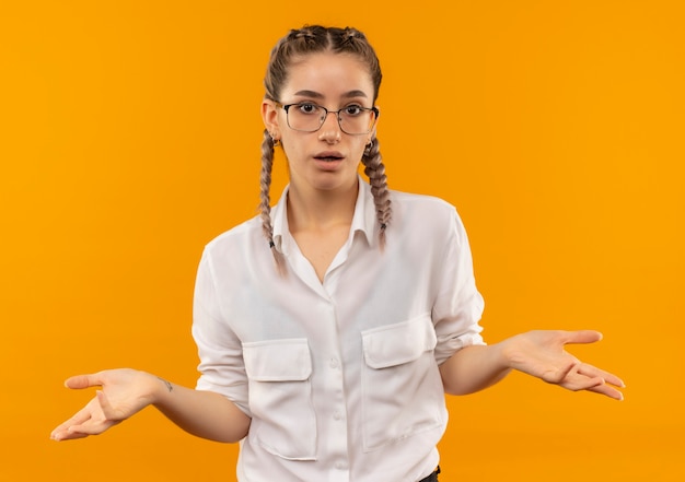 Foto gratuita ragazza giovane studente con gli occhiali con le trecce in camicia bianca guardando in avanti confuso e incerto diffondere le braccia ai lati senza risposta in piedi sopra il muro arancione