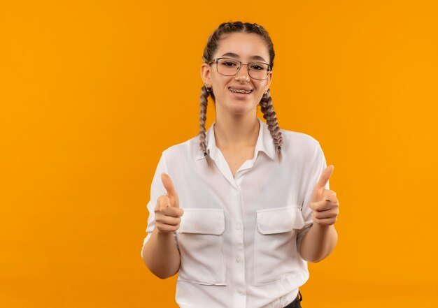 Молодая студентка в очках с косичками в белой рубашке смотрит вперед, весело улыбаясь, показывает палец вверх, стоя над оранжевой стеной