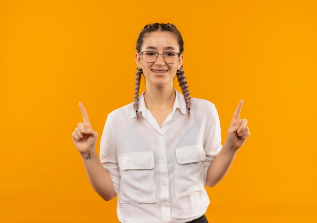 Young student girl in glasses with pigtails in white shirt looking to the front smiling pointing with index fingers up standing over orange wall