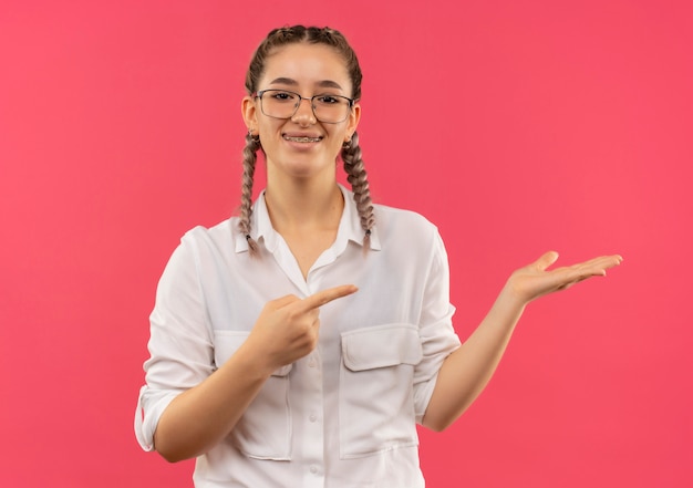 Молодая студентка в очках с косичками в белой рубашке смотрит вперед, улыбаясь, представляя что-то рукой, указывающей пальцем в сторону, стоящую над розовой стеной