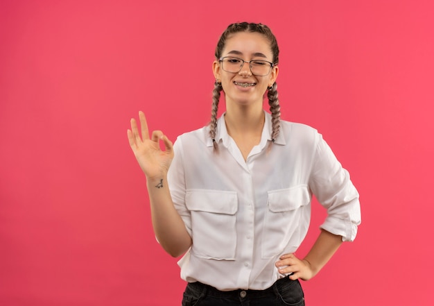Young student girl in glasses with pigtails in white shirt looking to the front smiling and winking showing ok sign standing over pink wall