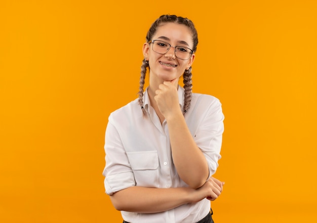 Молодая студентка в очках с косичками в белой рубашке смотрит вперед с уверенной улыбкой, стоя над оранжевой стеной