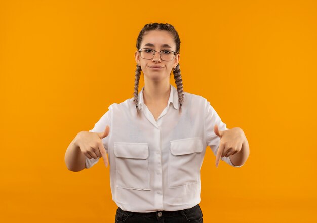 Молодая студентка в очках с косичками в белой рубашке смотрит вперед со скептическим выражением лица, указывая пальцами вниз, стоя над оранжевой стеной