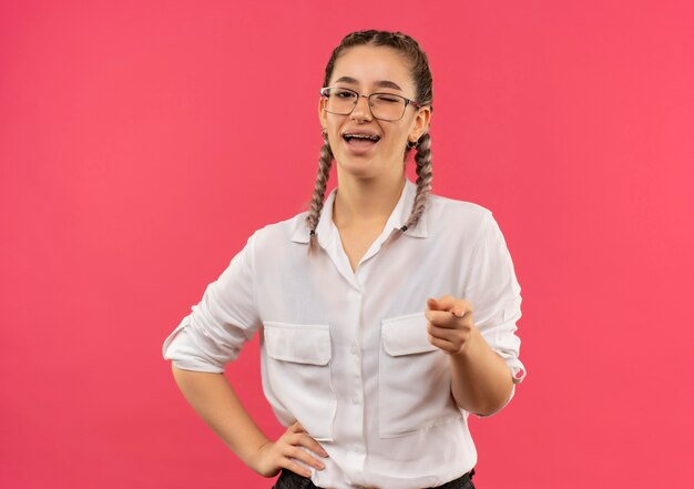 Young student girl in glasses with pigtails in white shirt pointing with index finger to the front smiling broadly happy and positive standing over pink wall