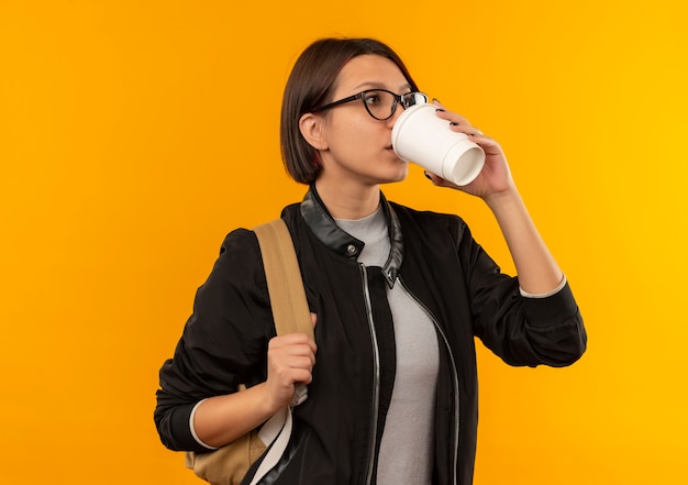 Молодая студентка в очках и с сумкой на спине пьет кофе из пластиковой кофейной чашки, глядя в сторону, изолированную на оранжевом фоне