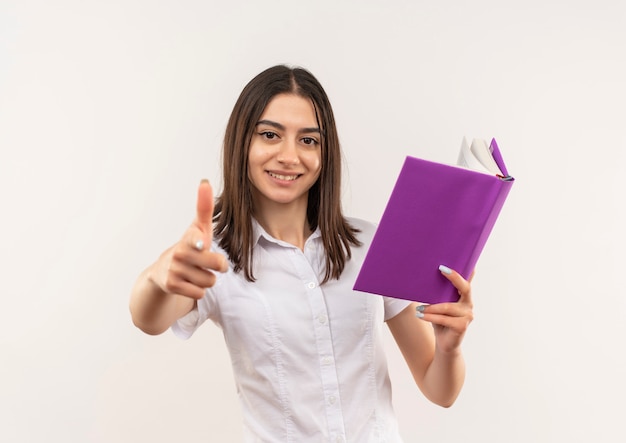 Foto gratuita ragazza giovane studente in camicia bianca che tiene libro aperto che mostra i pollici in su sorridente in piedi sopra il muro bianco