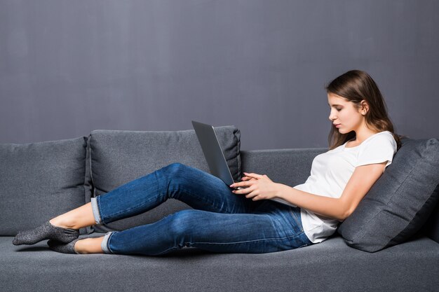 Young student girl in white t-shirt and blue jeans works on her laptop computer laying on grey coach sofa in front of grey wall