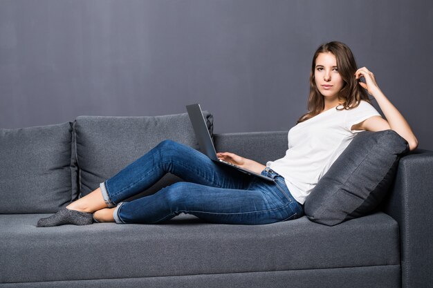 Young student girl in white t-shirt and blue jeans works on her laptop computer laying on grey coach sofa in front of grey wall