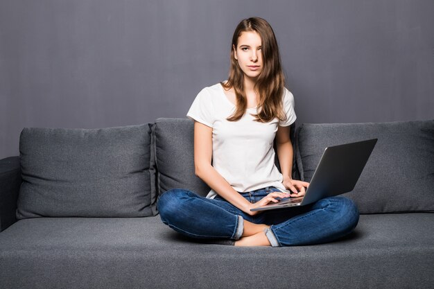 Young student girl in white t-shirt and blue jeans works on her laptop computer sitting on grey coach sofa in front of grey wall