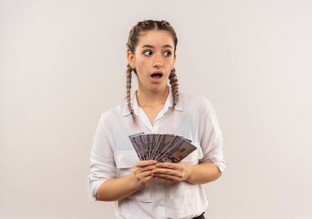 Foto gratuita ragazza del giovane studente con le trecce in camicia bianca che tiene contanti che osserva da parte in piedi sorpreso sopra il muro bianco