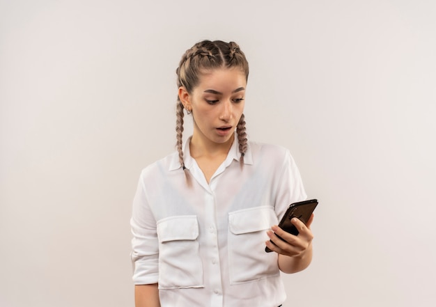 흰 벽에 서있는 혼란스러운 표정으로 그녀의 휴대폰의 화면을보고 흰 셔츠에 땋은 젊은 학생 소녀
