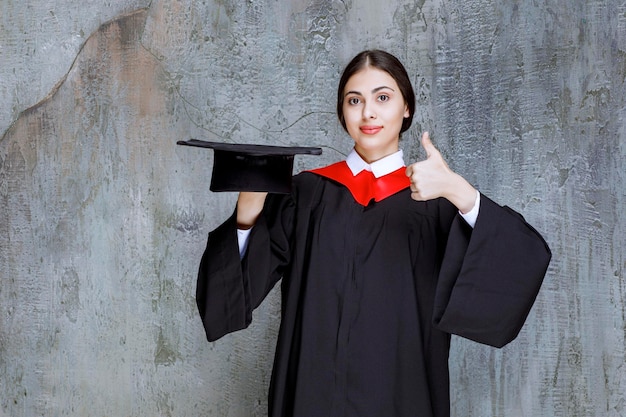 Молодой студент в платье держит шляпу и показывает палец вверх. Фото высокого качества
