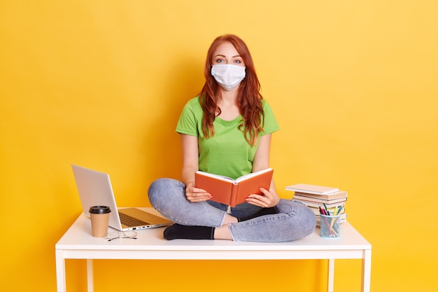 Young student in medical mask studying at home while quarantine, bored of distance learning, sitting with crossed legs on white table with book in hands.