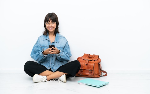Молодая студентка, сидящая на полу, изолированная на белом фоне, отправляет сообщение с мобильного телефона