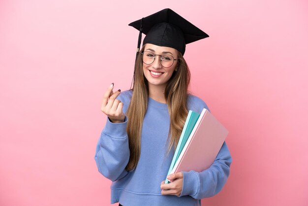Молодая студентка в шляпе выпускника на розовом фоне делает денежный жест