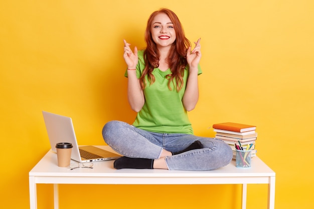 Young student woman in workplace with laptop and books, sitting with fingers crossing, wishes best, sitting with crossed legs on white table, looks smiling directly at camera.