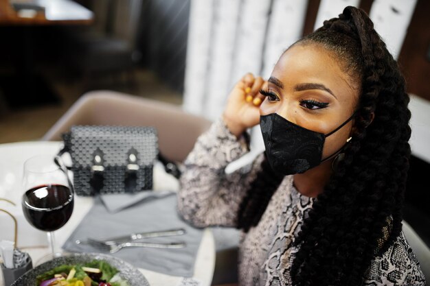 Young stylish african american woman wearing black face mask sitting in restaurant enjoying healthy food with wine