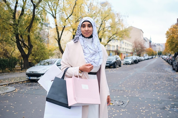 街の通りで買い物袋とカメラで自信を持って見ているヒジャーブの若いスタイリッシュなアラビア語の女性