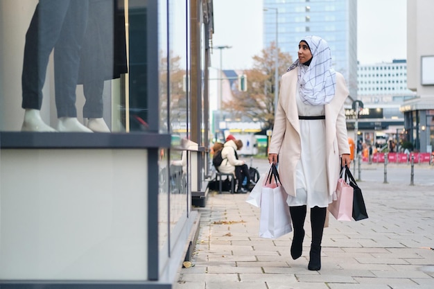 히잡을 쓴 젊고 세련된 아랍 여성이 쇼핑백을 들고 도시 거리를 꿈결같이 걷고 있다