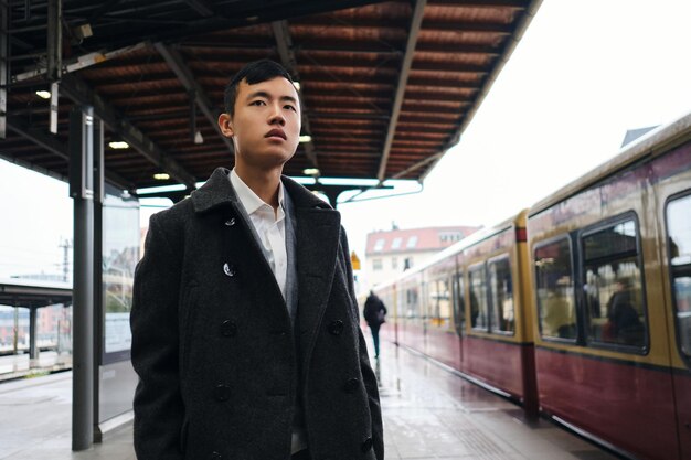 Young stylish Asian businessman intently looking away while waiting train on subway platform