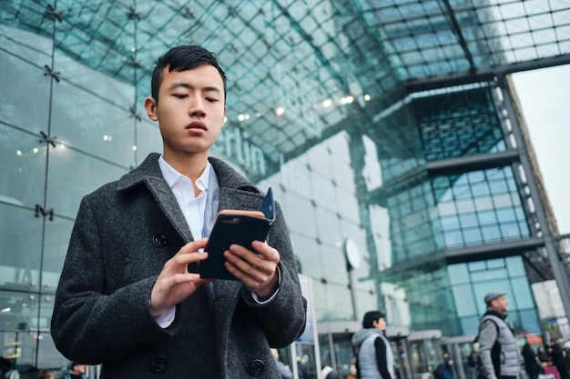 Giovane uomo d'affari asiatico alla moda che utilizza intensamente lo smartphone sulla strada vicino all'aeroporto