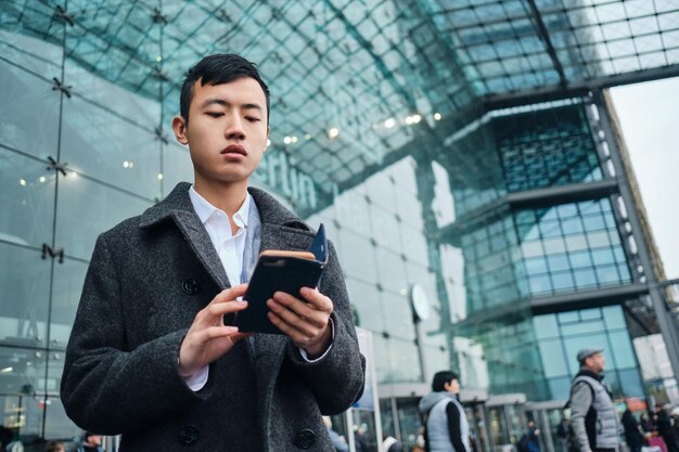Young stylish Asian businessman intently using smartphone on street near airport