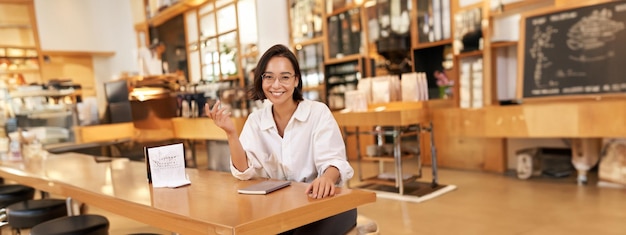 Молодая стильная азиатская женщина, владелец бизнеса в очках, сидит в кафе с ноутбуком, улыбается камере